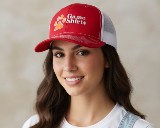 A smiling woman wearing a red and white trucker hat with a paw print and the words 'Game Shirts' on the front, paired with a white t-shirt and light blue denim overalls, set against a neutral beige background.