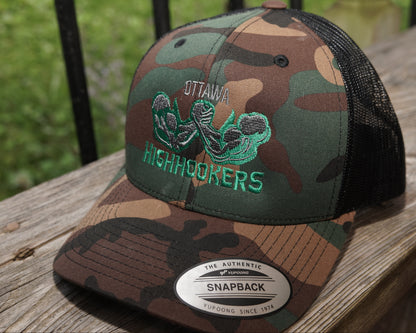 A camouflage snapback trucker hat with black mesh back panels, featuring a green embroidered 'Ottawa Highhookers' logo on the front. The hat is resting on a weathered wooden deck railing with a blurred green outdoor background.