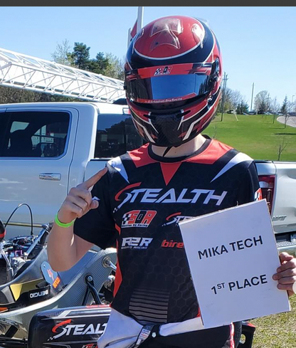 Person in racing gear holding a '1st Place' sign with a vehicle and track in the background
